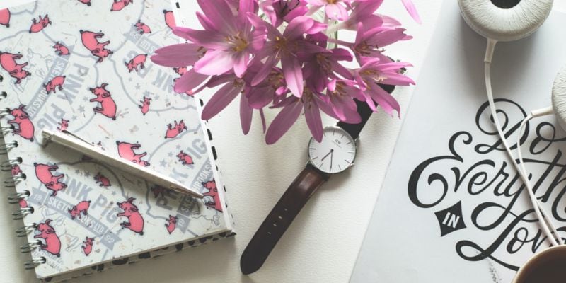 Top side view of black & pink stationary laid out on white surface A top view of a notebook, pencil, watch & writing in pink, black & white