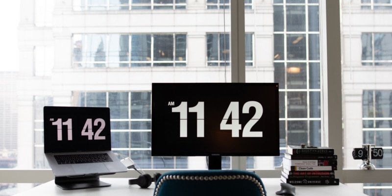 Home Office Desk With White Numbered Clock On Both A Desktop Computer Laptop Screen