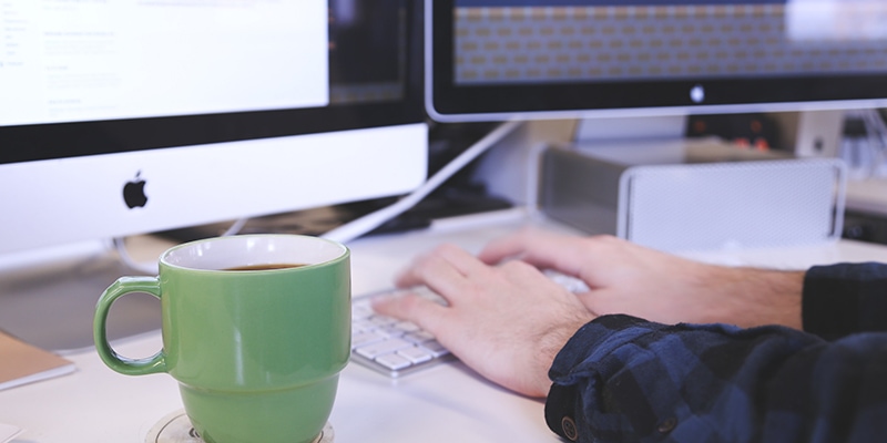Shot of mans arms typing on mac keyboard, with 2 screens in the background and green coffee mug infront