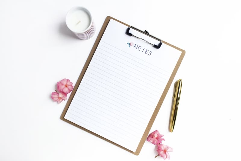 Lined Sheet Of Paper Titled Notes On A Clipboard With Pink Flowers A Gold Pen And A White Candle