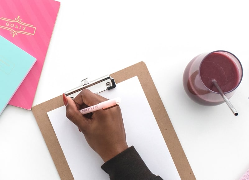 Woman Writing On Paper Against Clipboard With Smoothie Drink And Bright Colored Notebooks